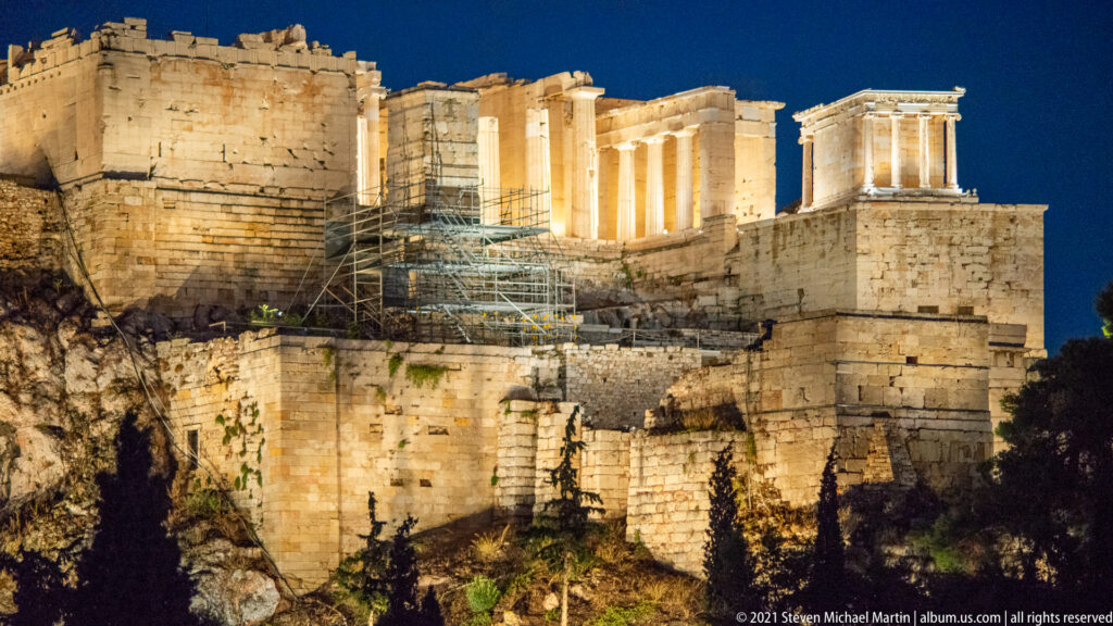 Illuminated Propylaia and Temple of Athena Nike above massive stone walls, rocky cliff, and scaffolding at night.
