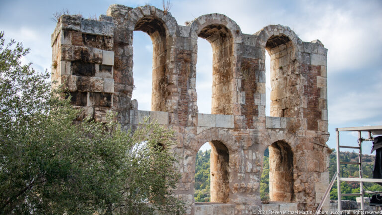 Tall arched openings in the stone exterior wall of the Odeon of Herodes Atticus, partly screened by an olive tree.