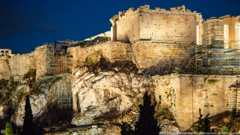 Night close view of illuminated Acropolis masonry rising from a jagged rock face with extensive scaffolding.
