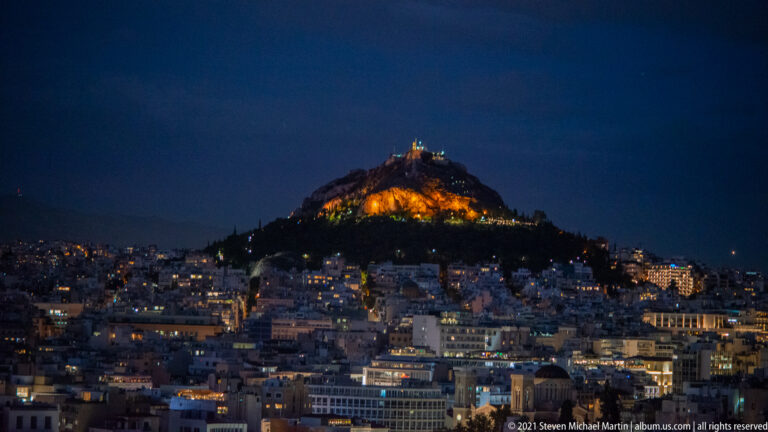Night view of Mount Lycabettus glowing above dense Athens, with a small summit building silhouetted at the top.