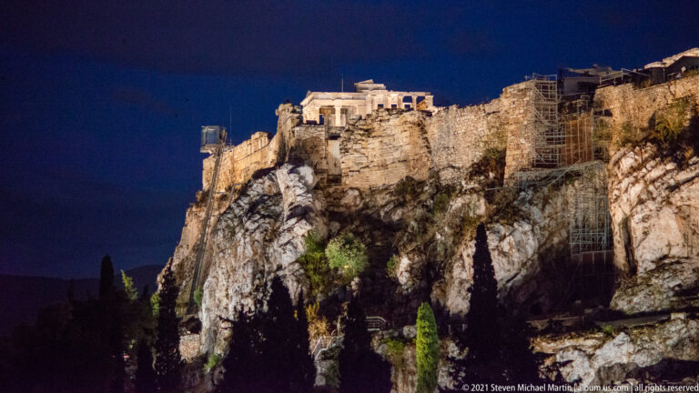 Night view of illuminated Acropolis walls above a steep pale limestone cliff with scaffolding and dark trees.