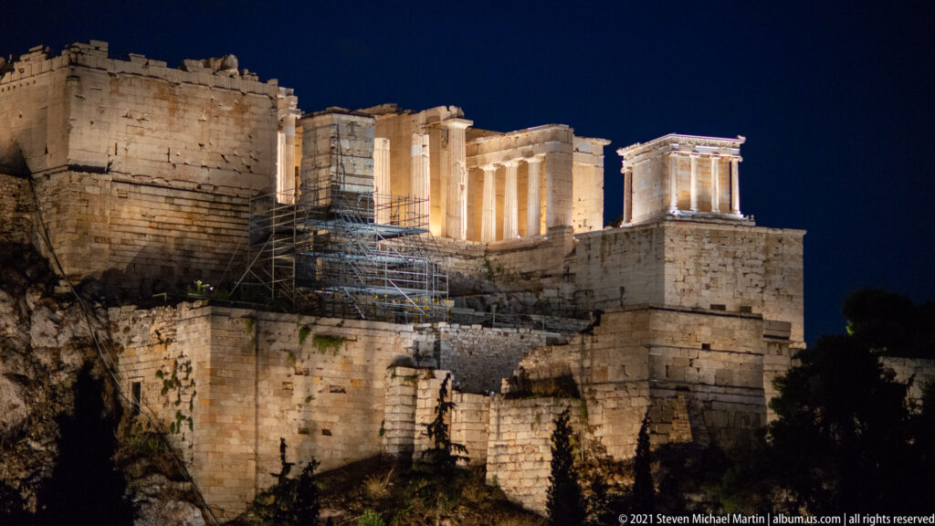 Close night view of the illuminated Propylaia and Temple of Athena Nike above walls and scaffolding.