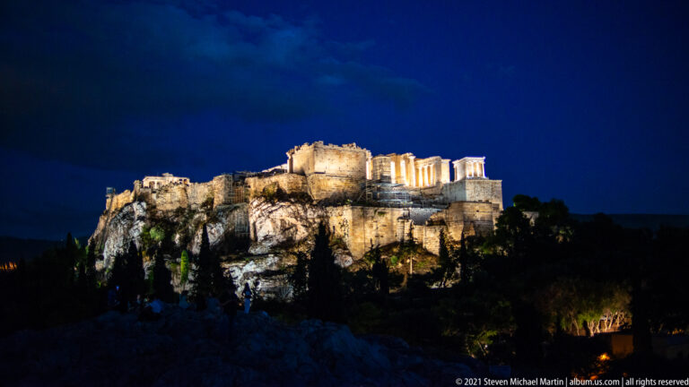 Wide night view of the illuminated western Acropolis ridge, monuments, walls, and rocky escarpment.