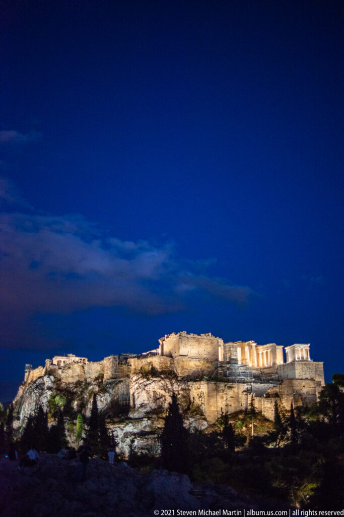 Distant panoramic night profile of the illuminated Acropolis across a dark foreground beneath a blue sky.