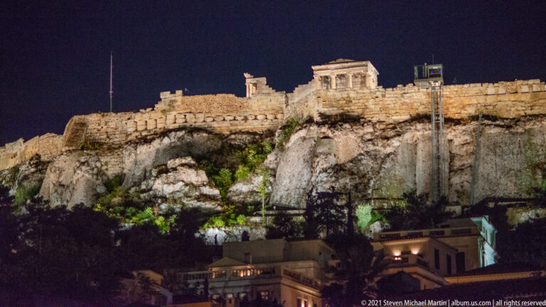 Illuminated Temple of Athena Nike above the Acropolis wall and rocky cliff, with city buildings below at night.