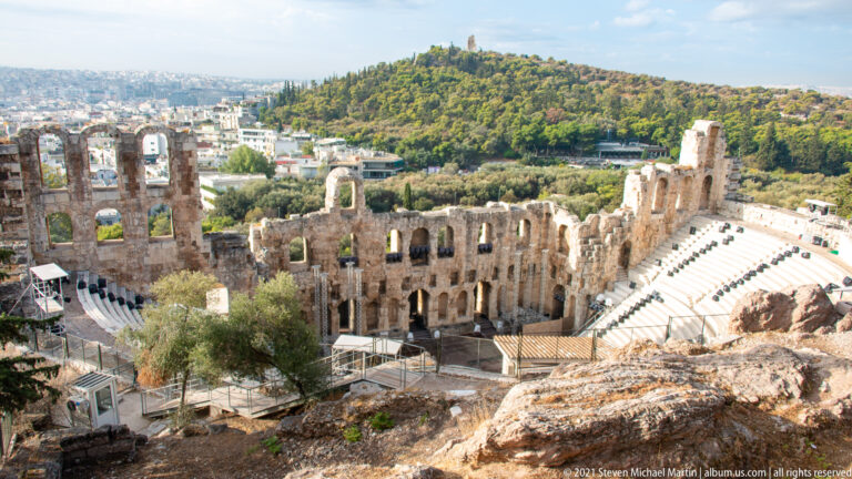 High daytime view of the Odeon of Herodes Atticus with curved seating, arched stage wall, and green hill beyond.