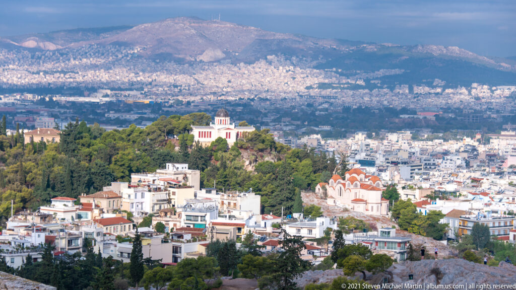 Athens cityscape beyond a wooded hill with churches and low buildings, seen from the Acropolis.