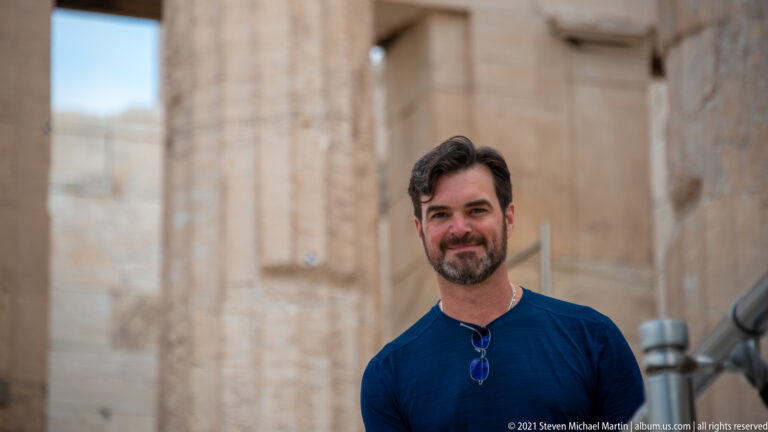 Male visitor in a dark blue shirt standing before softly blurred pale marble columns and walls on the Acropolis.