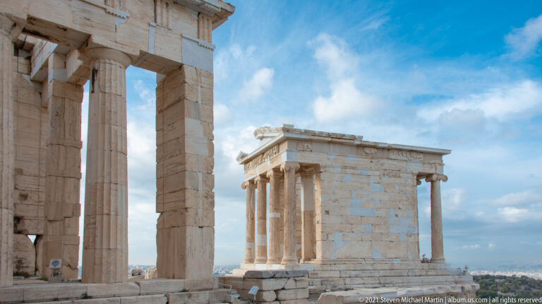 Temple of Athena Nike viewed between tall columns of the Propylaea against a blue, clouded sky.