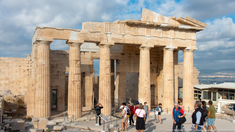 Visitors walking through the tall Doric columns and marble entrance of the Propylaea on the Acropolis.
