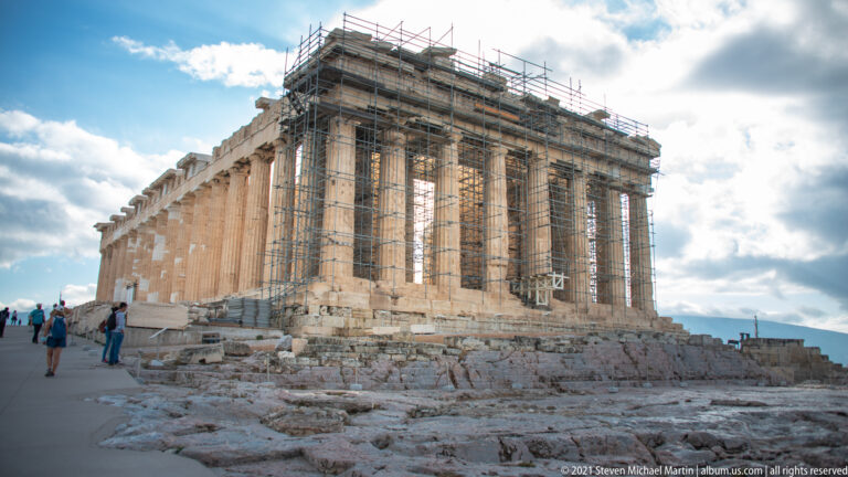 Parthenon with tall Doric columns and extensive scaffolding, viewed across rocky foundations on the Acropolis summit.
