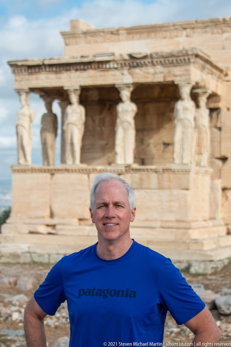 Male visitor in a blue shirt standing before the blurred Porch of the Caryatids at the Erechtheion.