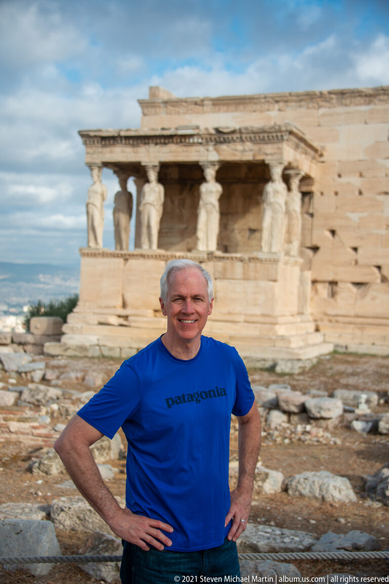Male visitor in a blue shirt standing on the Acropolis with the Erechtheion and Caryatid porch blurred behind.