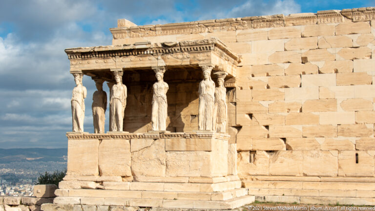 Porch of the Caryatids projecting from the Erechtheion, with six female supports and the Athens skyline beyond.