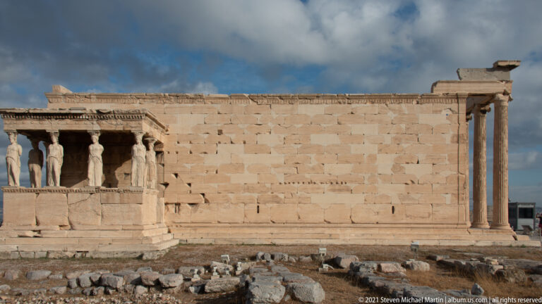 Side view of the Erechtheion with the Caryatid porch at left, long marble wall, and columned corner at right.