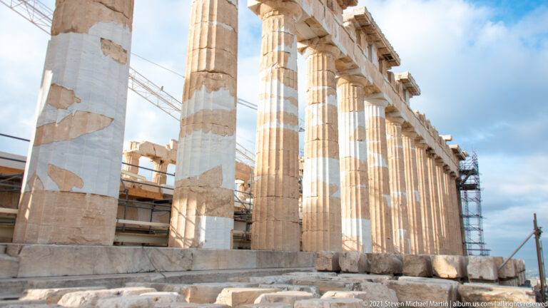 Oblique close view along the Parthenon’s fluted Doric columns, with pale restoration patches and scaffolding at the far end.