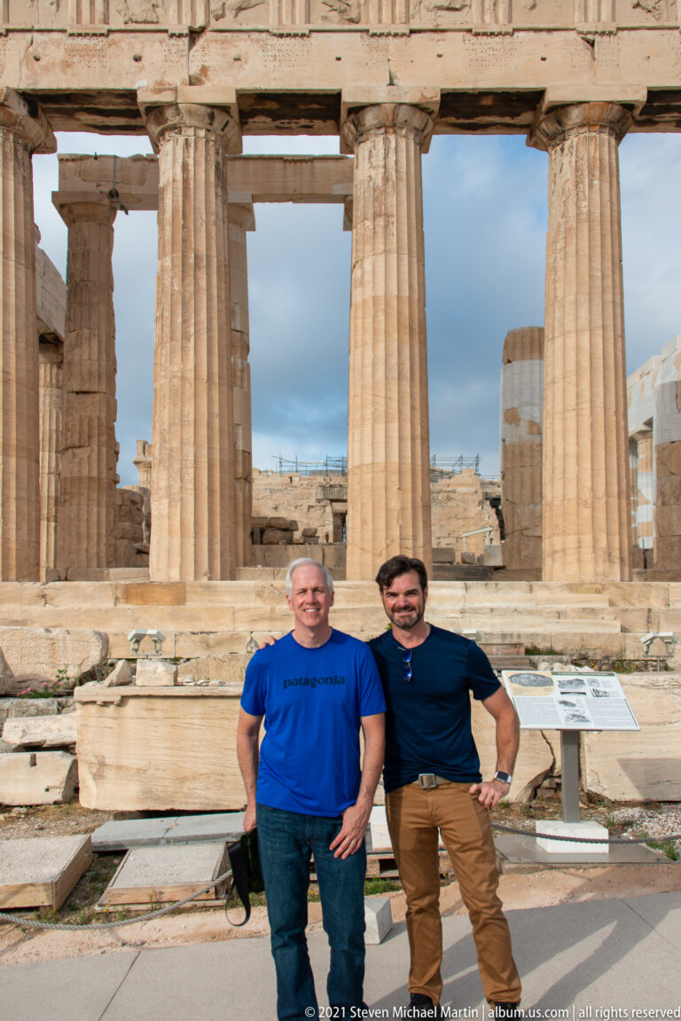 Two men standing in front of the Parthenon, with Doric columns and archaeological stonework behind them.