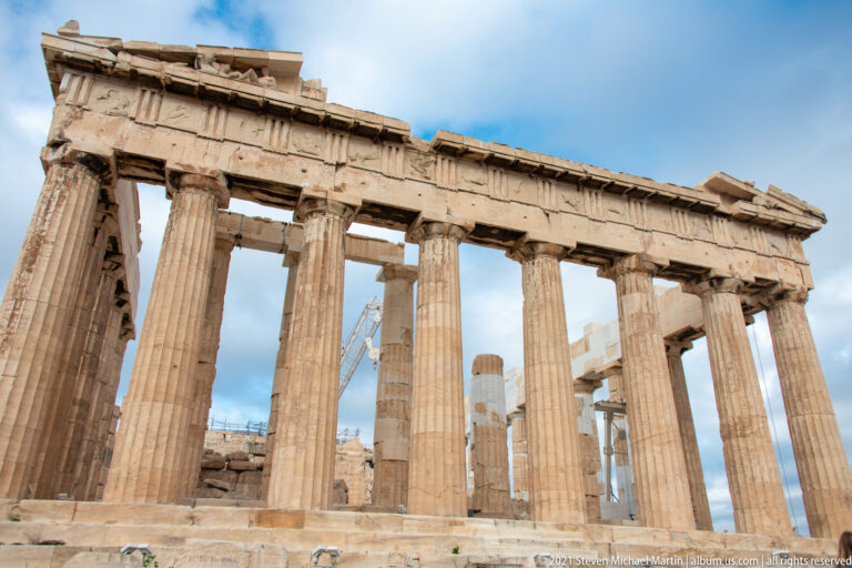 Low-angle corner view of the Parthenon showing intersecting rows of Doric columns against blue and cloudy sky.