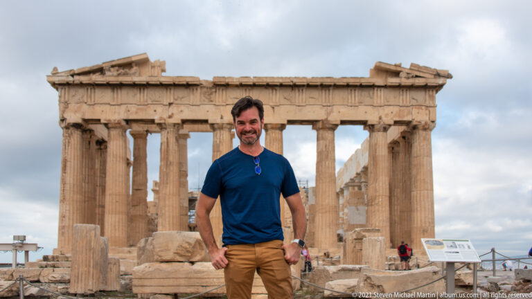 Man standing in front of the Parthenon, with its Doric columns, damaged pediment, and scattered ancient stone visible behind.