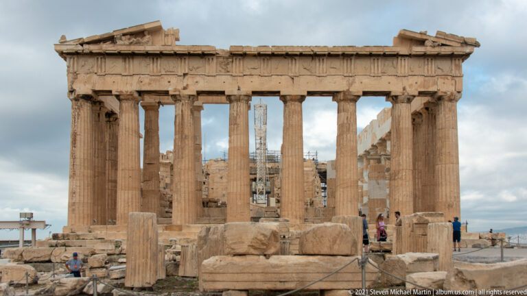 Frontal view of the Parthenon with Doric columns, broken pediment, scattered marble blocks, and visitors in the foreground.