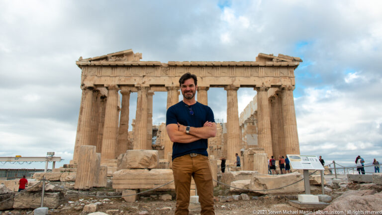 Man with arms crossed standing in front of the Parthenon, with columns, broken pediment, and visitors behind.