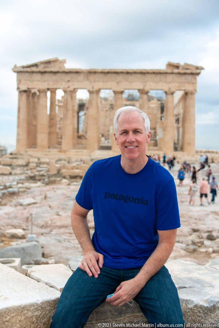 Seated man in a blue shirt with the Parthenon softly blurred across the background and other visitors on the Acropolis.