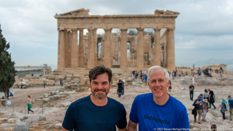 Two men standing together on the Acropolis with the Parthenon and many visitors behind them.