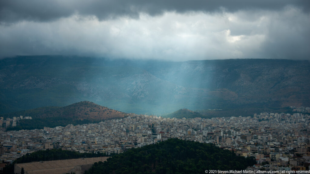 Dark storm clouds over Athens with a bright vertical rain shaft or sunlit shower descending onto the city below.
