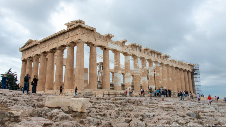 Oblique view of the Parthenon across rough Acropolis rock, with long rows of Doric columns, visitors, and distant scaffolding.