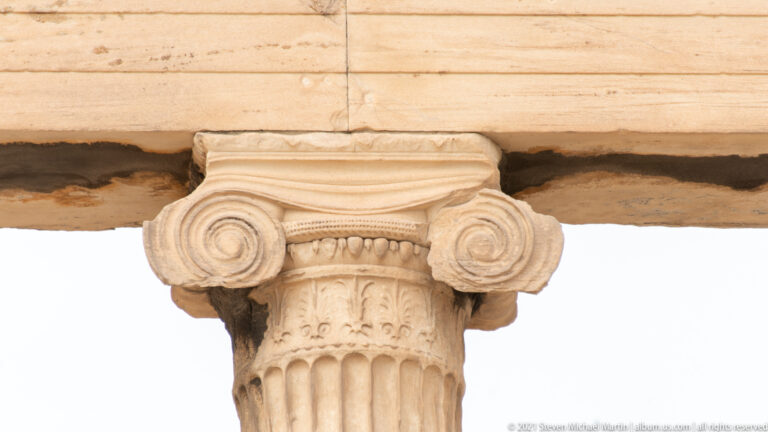 Close-up of an Ionic column capital with large spiral volutes, carved ornament, and the top of a fluted marble shaft.