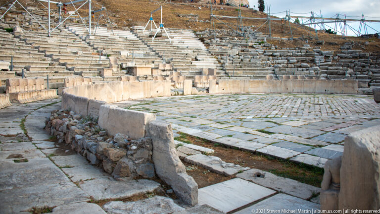 Paved orchestra and lower marble seating of the Theatre of Dionysus, with stairs and conservation scaffolding behind.