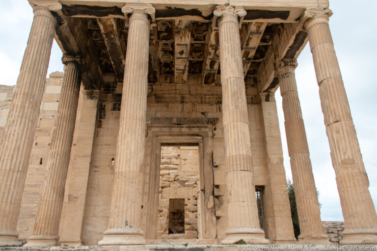 View beneath the Erechtheion’s north porch with Ionic columns, coffered ceiling, and a recessed stone doorway centered at the back.