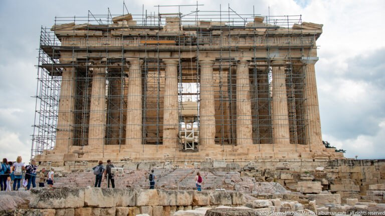 Wide frontal view of the Parthenon under heavy scaffolding, with visitors at the base and Doric columns visible behind the framework.
