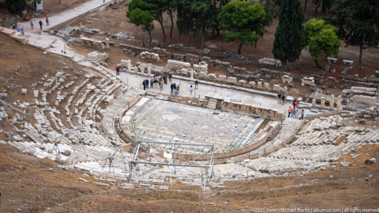 High-angle view of the Theatre of Dionysus with semicircular stone seating, orchestra area, visitors, and trees around the ancient theater.