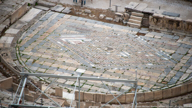 Close high-angle view of the Theatre of Dionysus orchestra, showing curved paving, patterned center, stage remains, and protective railings.