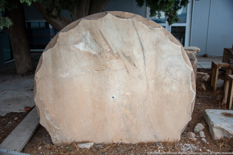 Large circular weathered marble fragment standing upright beside conservation shelving and scattered stone pieces.