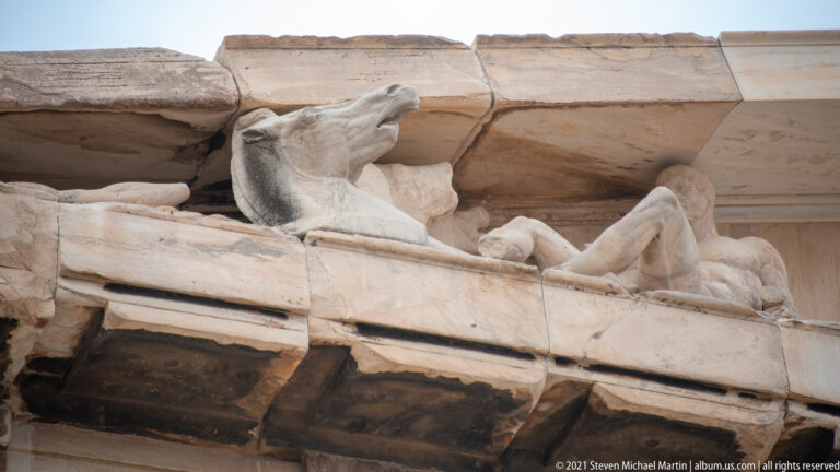 Close view of a marble horse head and reclining human figure beneath a projecting stone cornice.