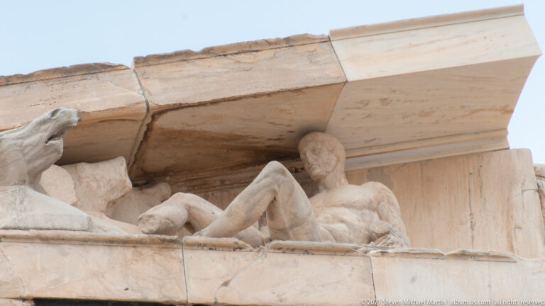 Weathered reclining marble figure beneath a heavy cornice, surrounded by restored stonework and fragmentary sculpture.