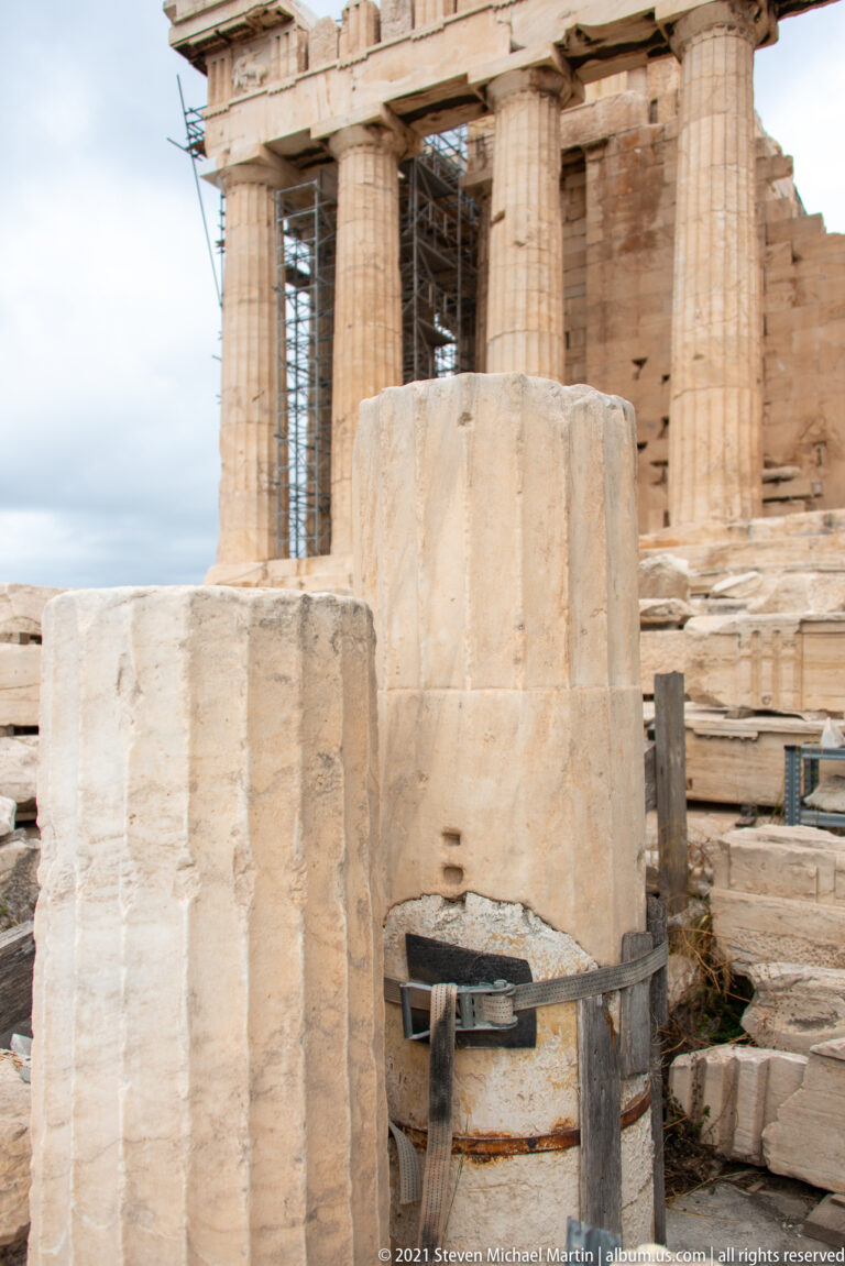Large fluted marble column drums and stone blocks in front of the Parthenon colonnade and scaffolding.