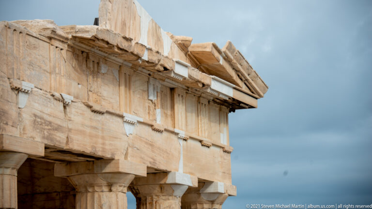 Close view of an ancient marble wall and projecting entablature with numerous lighter replacement blocks.