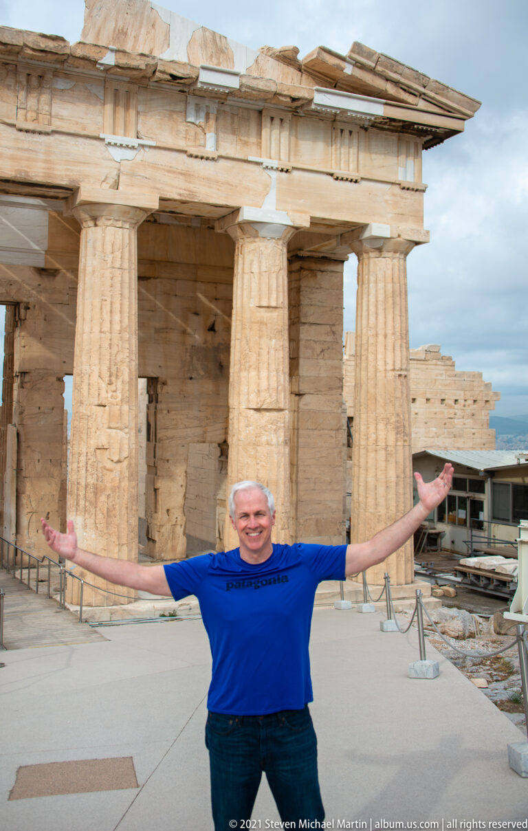 Male visitor with arms outstretched in front of large fluted columns and marble gateway architecture at the Propylaia.