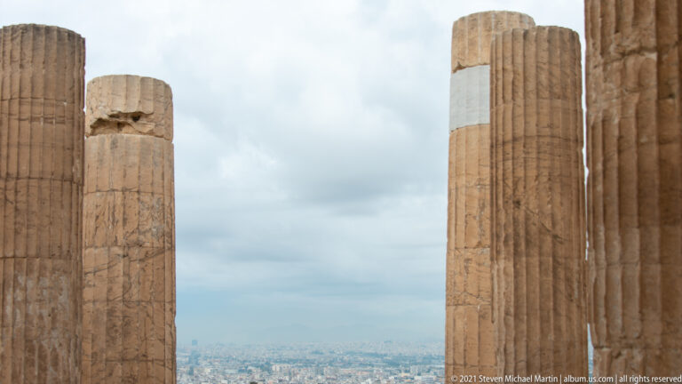 Tall weathered fluted stone columns with white repair bands, framing a distant view of Athens.