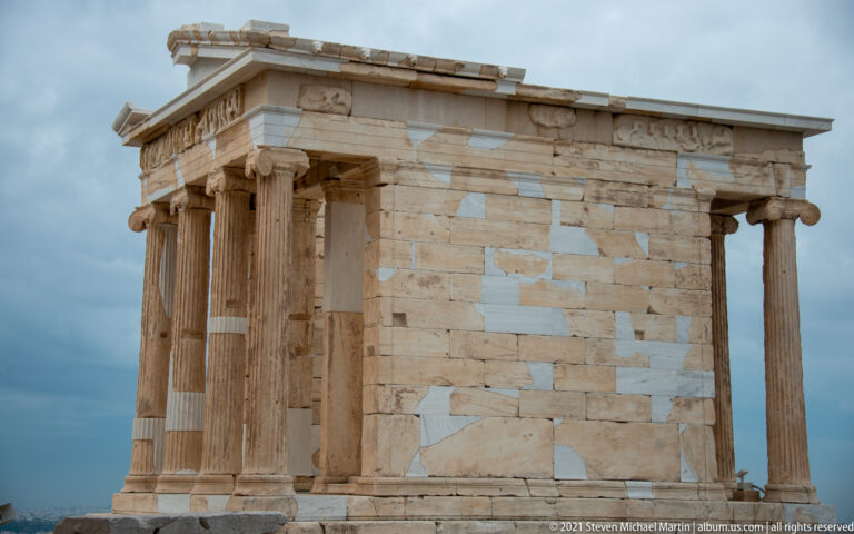 Side view of the small Ionic Temple of Athena Nike with restored marble blocks under a cloudy sky.