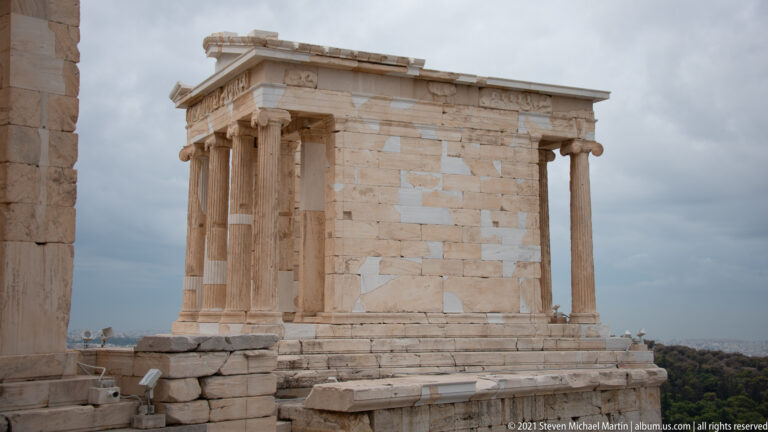 Small Ionic Temple of Athena Nike standing on a raised stone bastion beneath gray clouds.