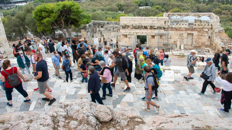 Crowd of visitors walking over marble paving beside low ancient stone walls on the Acropolis.