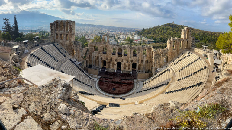 Wide elevated view of the Odeon of Herodes Atticus, showing curved seating, stage wall, city, and hills.