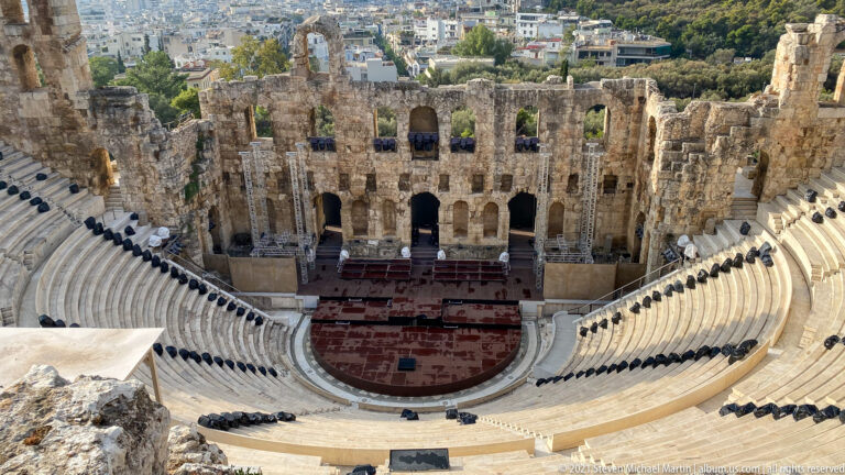 High view centered on the Odeon’s reddish orchestra, arched stage wall, and curved tiers of pale seating.