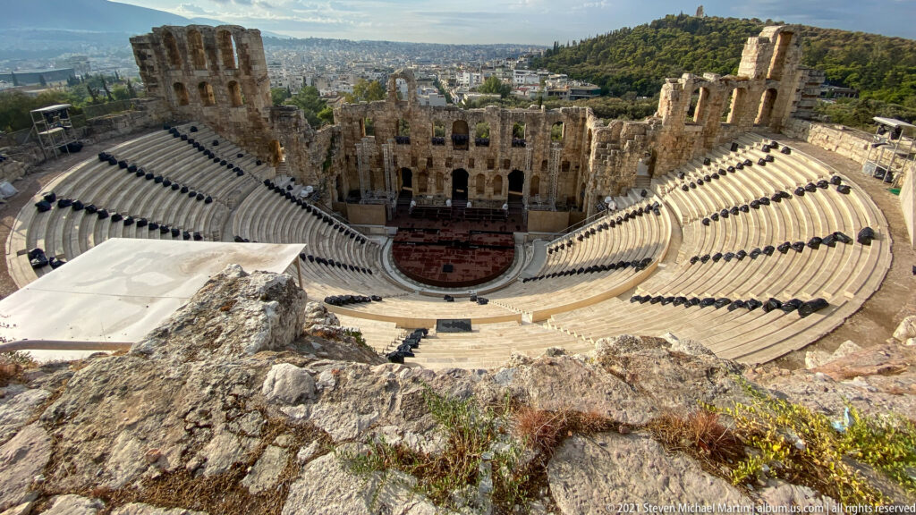 Wide view over rocky foreground to the Odeon of Herodes Atticus, Athens, and wooded hills.