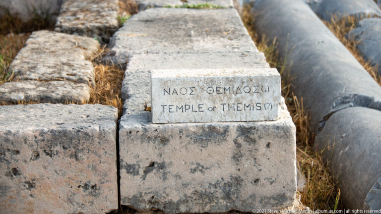 Small marble marker reading “Temple of Themis” on ancient stone blocks beside a fallen column shaft.