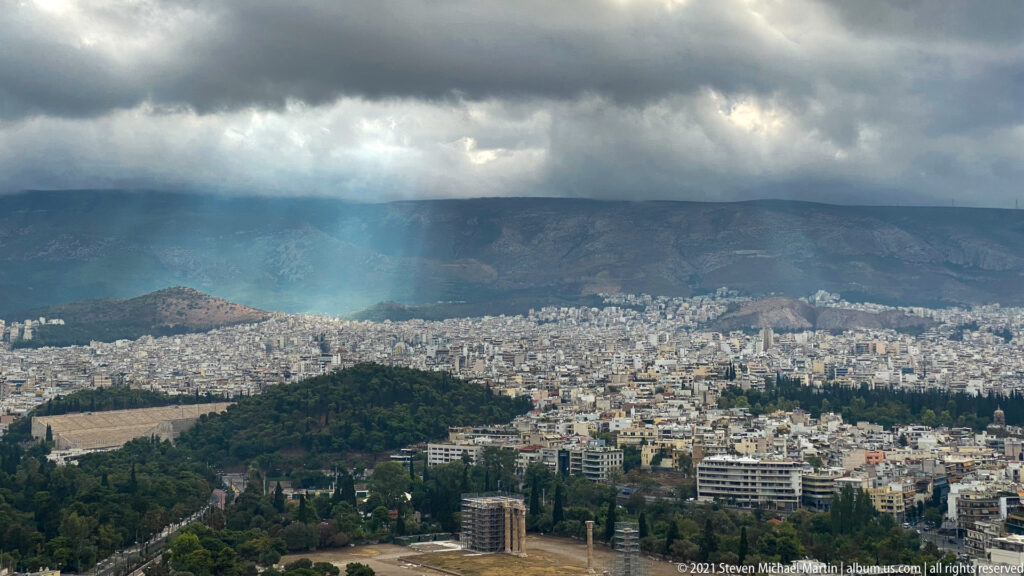 Athens cityscape beneath dark clouds, with sunbeams, a wooded hill, ancient remains, and mountains beyond.